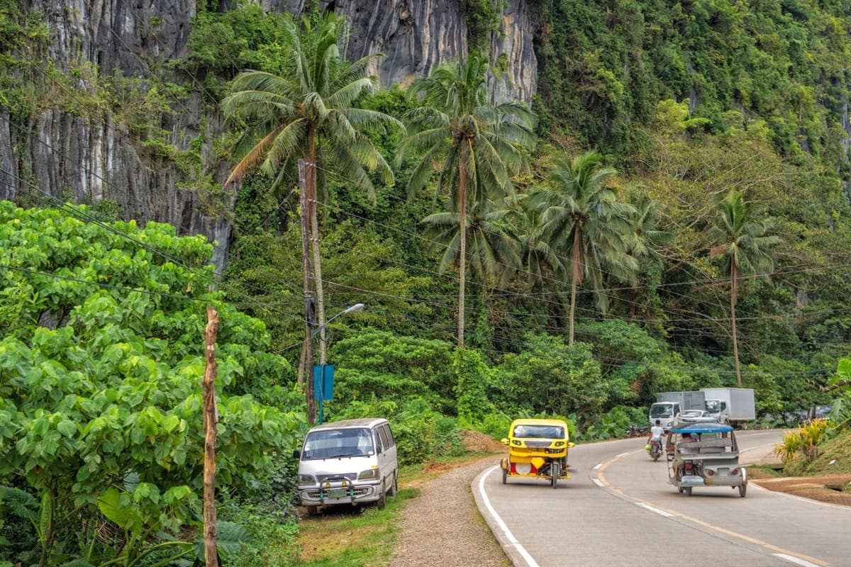 Yellow tricycle passing a parked white van on a road beneath limestone cliffs and palms