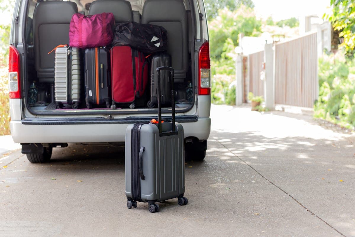 Suitcases and bags stacked in the open rear of a van, with a wheeled cabin case on the ground