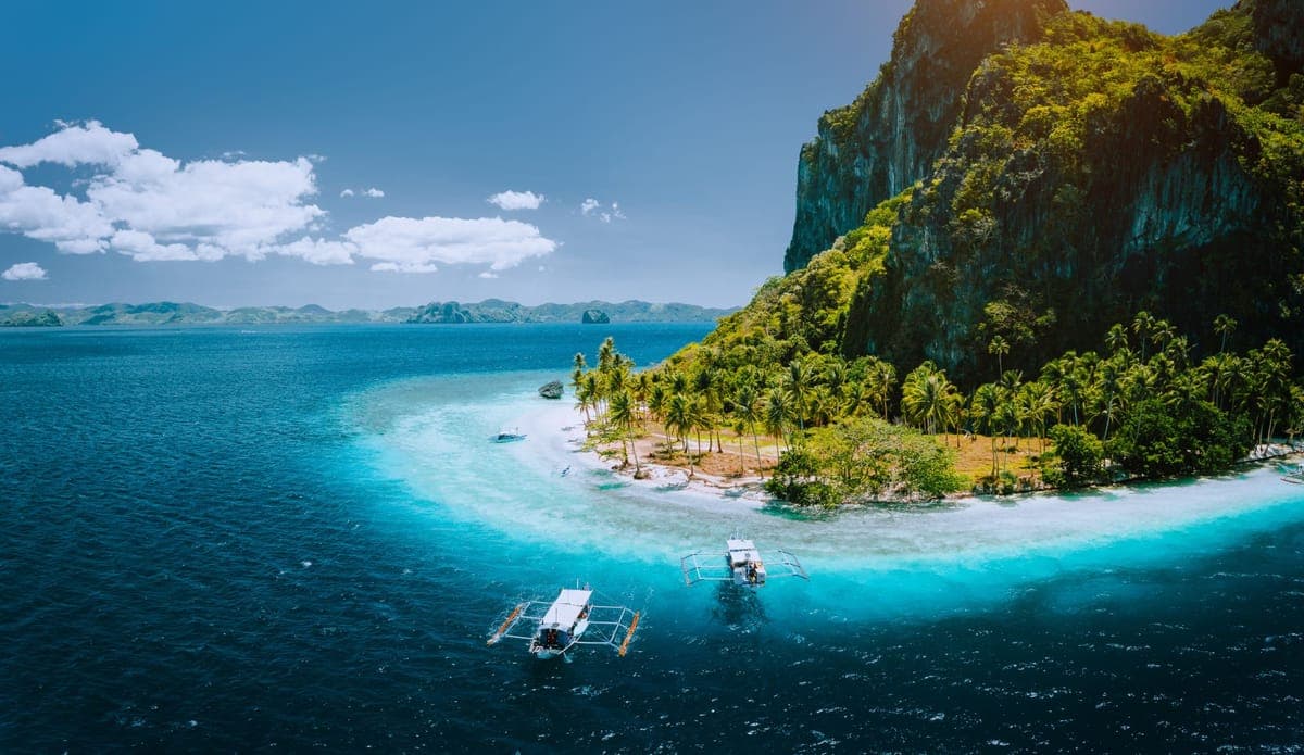 Outrigger boats on a turquoise lagoon beside a palm-fringed limestone island, seen from the air